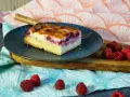 Ein Stück Himbeer Schmand Kuchen auf einem blauen Teller, daneben frische Himbeeren.
