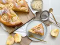 Ein köstlicher veganer Apfelkuchen, bestäubt mit Puderzucker, wird auf einem Holzbrett präsentiert. Daneben liegen frische Apfelstücke und eine Schale mit Mandelblättchen.