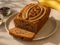 Ein saftiges Bananenbrot ohne Zucker, garniert mit Bananenscheiben und Schokostückchen. Daneben Schüsseln mit Nüssen und Schokodrops.