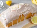 Saftiger zitronenkuchen in Kastenform, überzogen mit süßem Zuckerguss. Ein Zitronenstück liegt daneben.