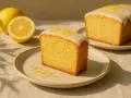 Ein Stück saftiger, veganer Zitronenkuchen mit Zuckerguss und Zitronenschale bestreut. Daneben liegen frische Zitronen.