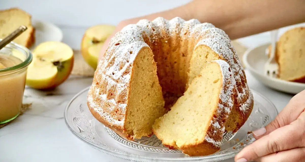 Ein saftiger Kuchen mit Apfelmus, bestäubt mit Puderzucker, daneben Äpfel und ein Glas Apfelmus.