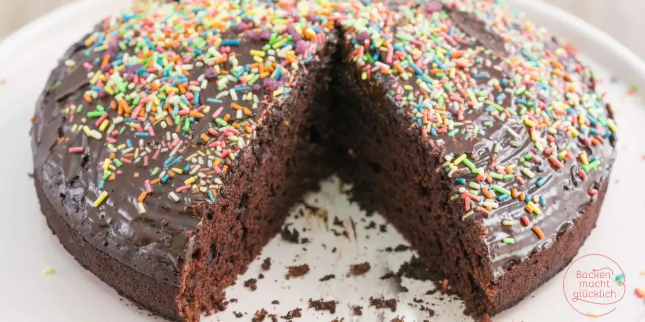 Ein saftiger, veganer Kuchen mit Schokoladenglasur und bunten Streuseln. Ein Stück fehlt, was Lust auf mehr macht.