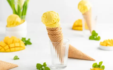 Zwei Kugeln cremiges Mango Eis in Waffelhörnchen, umgeben von frischen Mangostücken und Minze. Ein süßer Genuss!