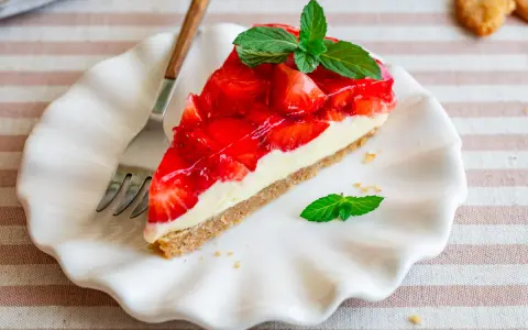 Ein Stück Erdbeerkuchen ohne Backen, mit frischen Erdbeeren und Minze garniert, auf einem weißen Teller.