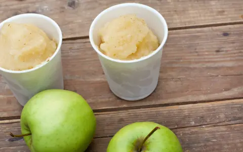 Zwei Becher mit erfrischendem Apfel Sorbet stehen neben zwei grünen Äpfeln auf einer Holzoberfläche.