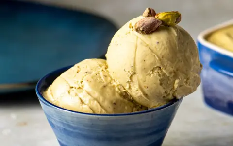 Zwei Kugeln cremiges, gelbes Eis mit Pistazienstückchen, garniert mit ganzen Pistazien, in einer blauen Schale.