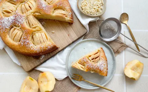 Ein köstlicher veganer Apfelkuchen, bestäubt mit Puderzucker, wird auf einem Holzbrett präsentiert. Daneben liegen frische Apfelstücke und eine Schale mit Mandelblättchen.