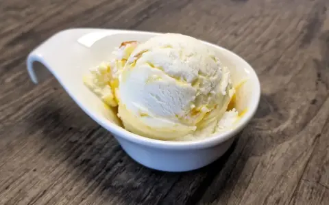 Ein Löffel cremiges Buttermilch-Eis, beträufelt mit Honig, in einer weißen Schale auf Holztisch.