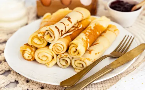 Ein Teller mit goldbraunen Quark Pfannkuchen, aufgerollt und gestapelt. Daneben liegen ein goldenes Messer und eine Gabel. Im Hintergrund eine Schale Marmelade.