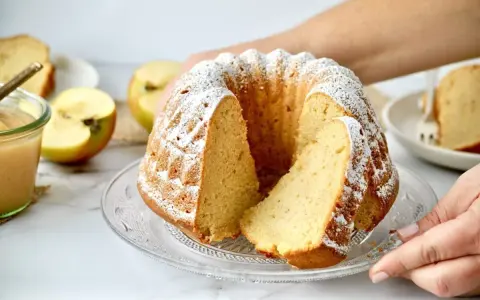 Ein saftiger Kuchen mit Apfelmus, bestäubt mit Puderzucker, daneben Äpfel und ein Glas Apfelmus.