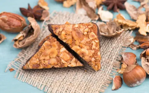 Zwei vegane Nussecken mit Schokoladenrand, bedeckt mit gehackten Nüssen, liegen auf einem Jutesack, umgeben von verschiedenen Nüssen und Sternanis.