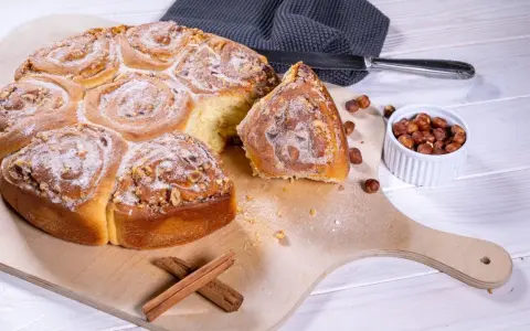 Ein köstliches Zimtschnecken Rezept: goldbraune, mit Nüssen und Puderzucker bestreute Schnecken auf einem Holzbrett, daneben Zimtstangen und Haselnüsse.