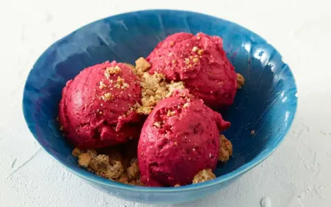 Drei Kugeln leuchtend pinkes Sauerrahm Eis, bestreut mit knusprigen Krümeln, in einer blauen Schale.