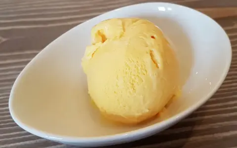 Ein Löffel cremiges, gelbes balla balla Eis in einer weißen Schale auf Holzhintergrund.