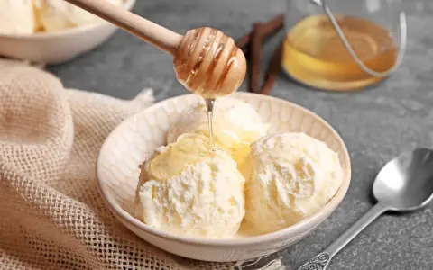 Schneeballgroße Kugeln Vanilleeis werden mit flüssigem Honig beträufelt. Ein Holzlöffel tropft goldenen Honig auf das cremige Eis.