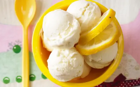 Erfrischendes Buttermilch Zitronen Eis in einer gelben Schale, garniert mit Zitronenscheiben. Ein gelber Löffel liegt daneben.