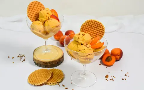 Zwei Gläser mit cremigem Aprikosen Eis, garniert mit frischen Aprikosenstücken und Waffeln. Ein sommerlicher Genuss!