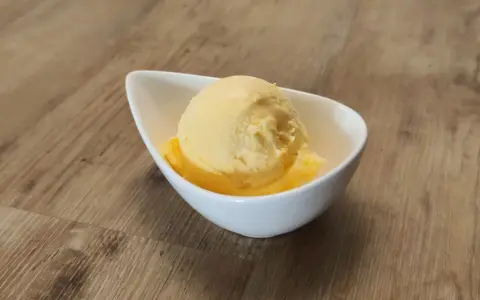 Ein Löffel cremiges Mandarinen Eis in einer weißen Schale auf Holzhintergrund.