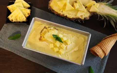 Erfrischendes Ananas Eis in einer Metallform, garniert mit Ananasstücken und Minze. Daneben liegen Ananasspalten und eine Waffel.