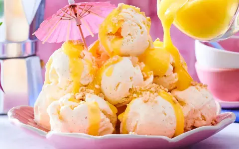 Ein Berg aus cremigem Vanilleeis, übergossen mit warmer Zitronensauce und bestreut mit Krümeln. Ein rosa Sonnenschirm sorgt für tropisches Flair.