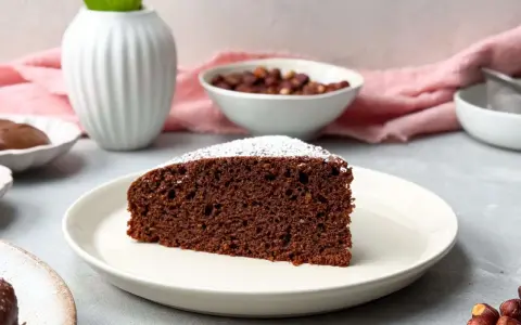 Ein saftiges Stück **Nutella Kuchen** mit Puderzucker bestäubt, daneben Nüsse und eine Vase.