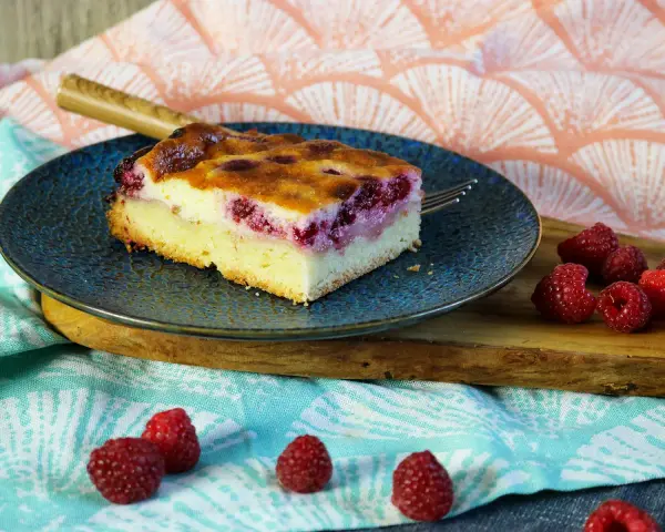 Ein Stück Himbeer Schmand Kuchen auf einem blauen Teller, daneben frische Himbeeren.