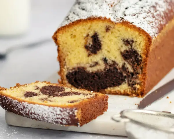 Ein saftiger Marmorkuchen in Kastenform, bestäubt mit Puderzucker. Eine Scheibe liegt daneben, bereit zum Genuss.
