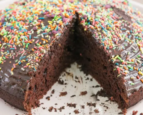 Ein saftiger, veganer Kuchen mit Schokoladenglasur und bunten Streuseln. Ein Stück fehlt, was Lust auf mehr macht.