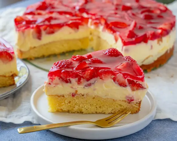 Ein saftiger Erdbeer Schmand Kuchen mit frischen Erdbeeren und rotem Tortenguss, ein Stück auf einem Teller mit einer goldenen Gabel.