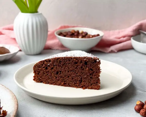 Ein saftiges Stück **Nutella Kuchen** mit Puderzucker bestäubt, daneben Nüsse und eine Vase.