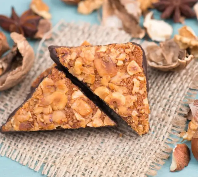 Zwei vegane Nussecken mit Schokoladenrand, bedeckt mit gehackten Nüssen, liegen auf einem Jutesack, umgeben von verschiedenen Nüssen und Sternanis.