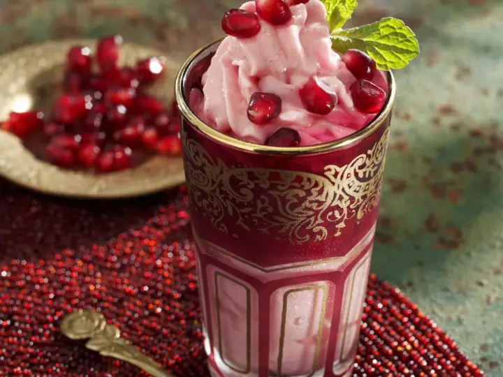 Ein Glas mit cremigem granatapfel eis, garniert mit frischen Granatapfelkernen und Minze. Daneben eine Schale mit Granatapfelkernen.