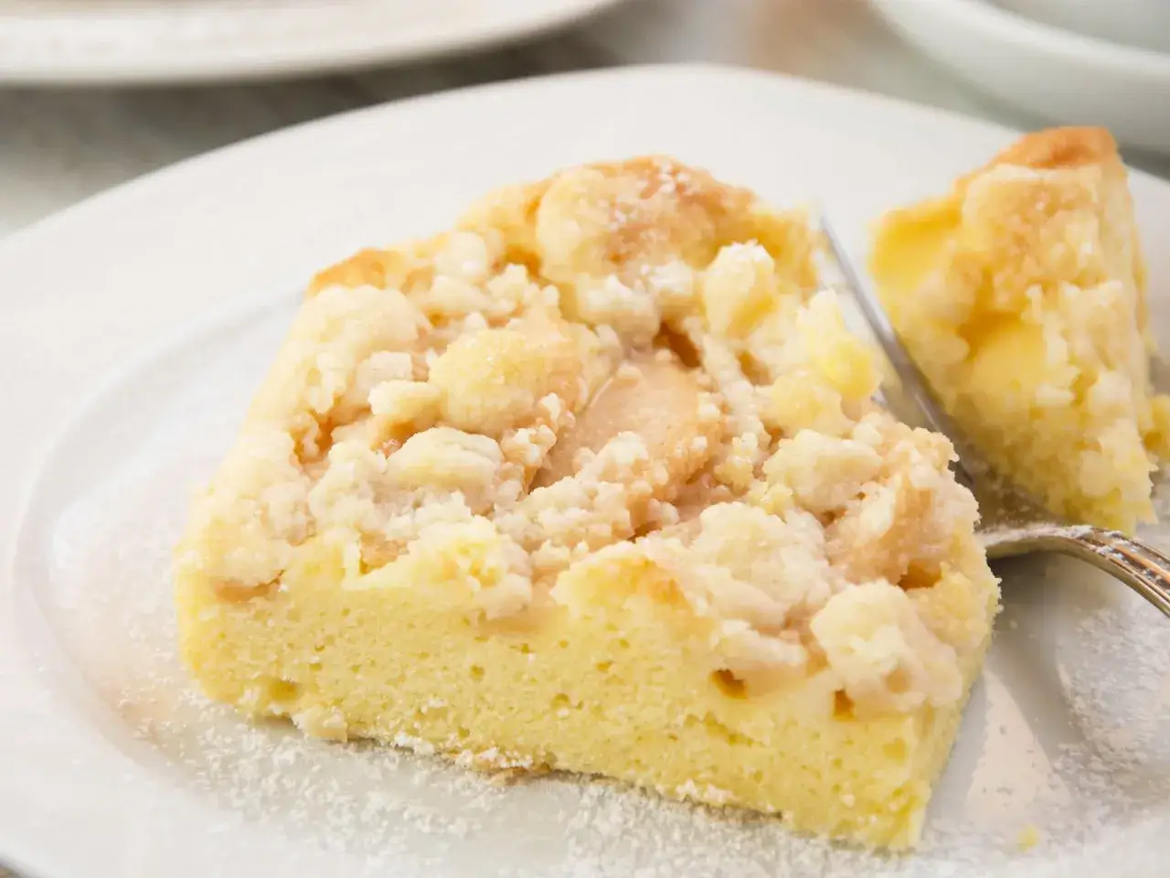 Saftiger Kuchen mit Apfelmus und Streuseln, bestäubt mit Puderzucker. Ein Stück ist bereits angeschnitten.