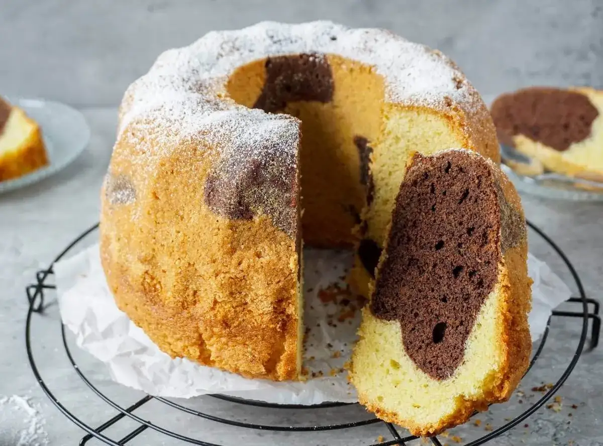 Ein saftiger Marmorkuchen mit Puderzucker bestäubt, ein Stück davon ist abgetrennt und zeigt die schöne Marmorierung.