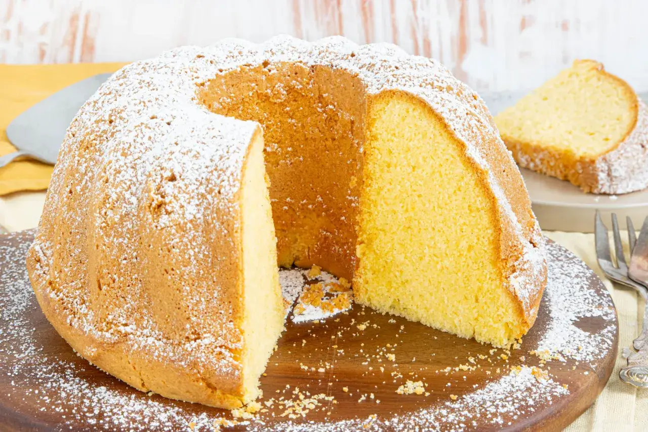 Ein saftiger vanille kuchen, bestäubt mit Puderzucker, liegt auf einem Holzbrett. Ein Stück ist bereits angeschnitten.