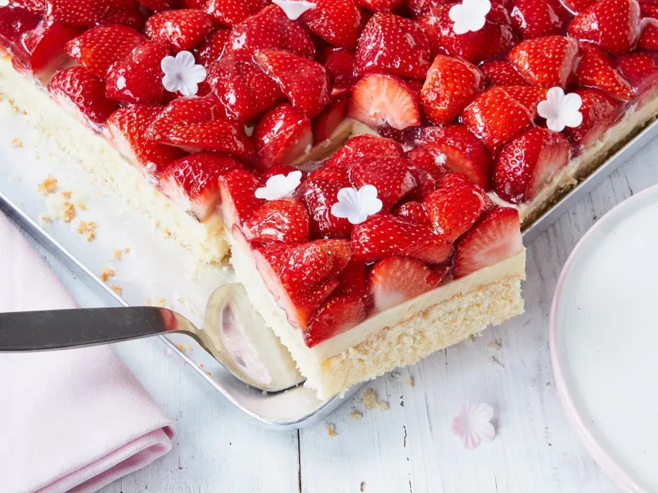 Ein saftiger Erdbeerkuchen vom Blech, bedeckt mit glänzenden Erdbeeren und kleinen weißen Blüten. Ein Stück wurde bereits entnommen.