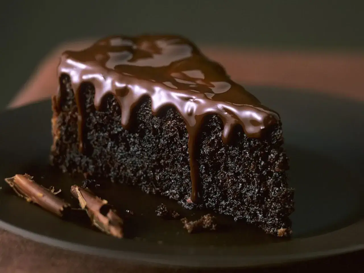 Ein Stück saftiger Schoko Kuchen mit glänzender Schokoladenglasur, die über den Rand tropft. Daneben liegen zwei Schokostückchen.