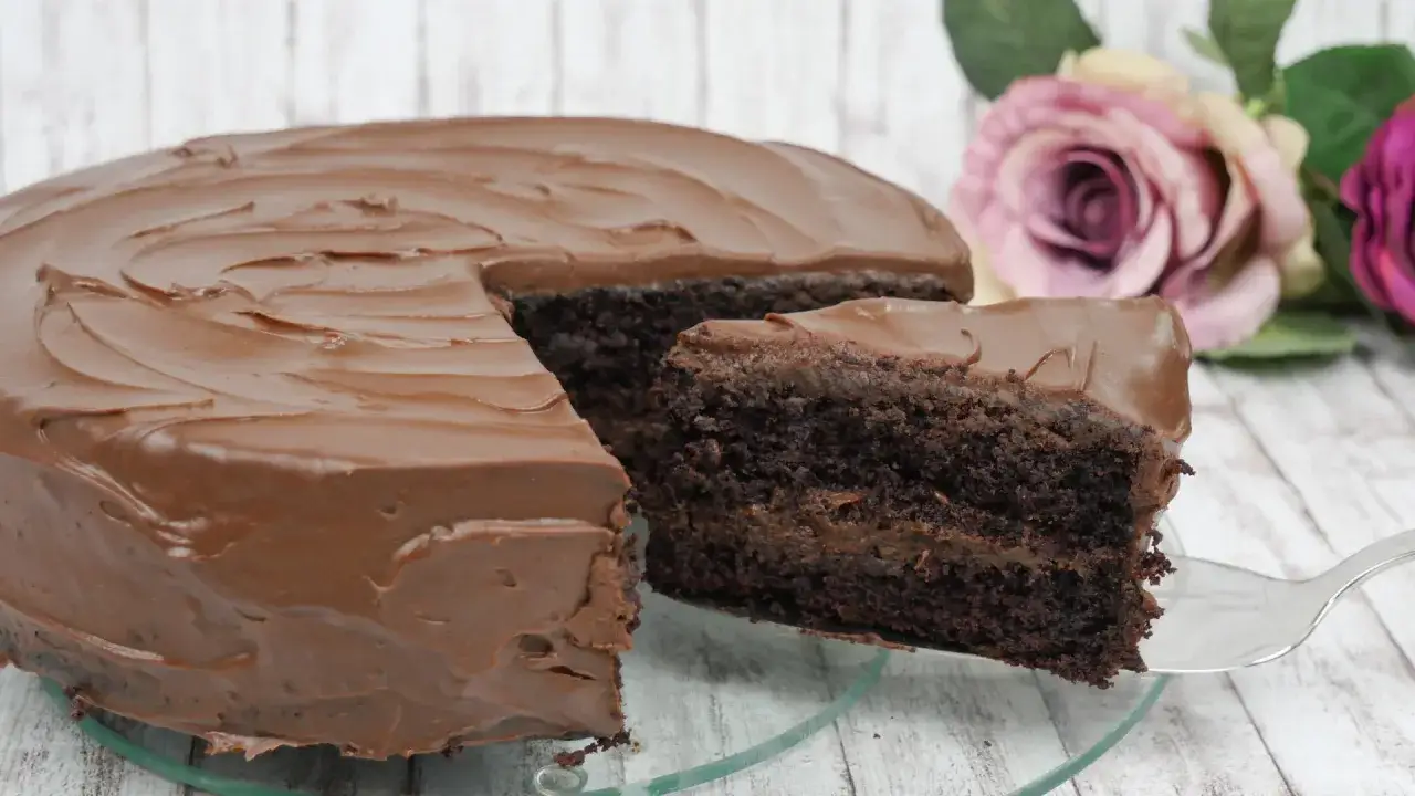 Ein saftiger schoko kuchen mit Schokoladenglasur, ein Stück wird gerade herausgehoben. Rosen im Hintergrund.