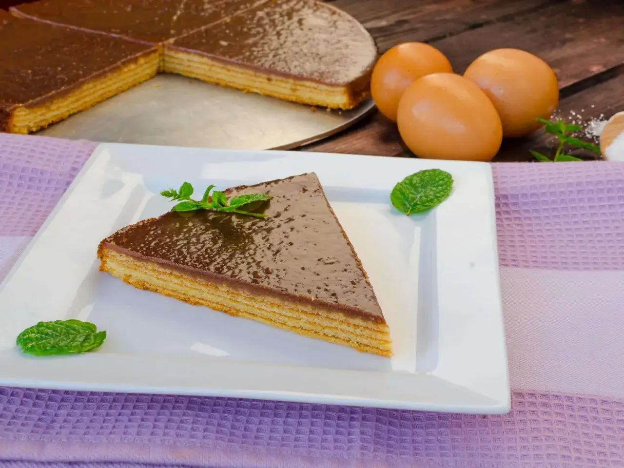 Ein Stück schokoladenglasierter Baumkuchen mit Minze, daneben Eier und Zutaten für ein Baumkuchen Rezept.