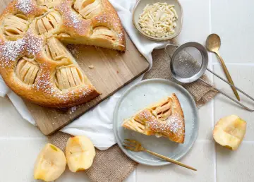 Ein köstlicher veganer Apfelkuchen, bestäubt mit Puderzucker, wird auf einem Holzbrett präsentiert. Daneben liegen frische Apfelstücke und eine Schale mit Mandelblättchen.