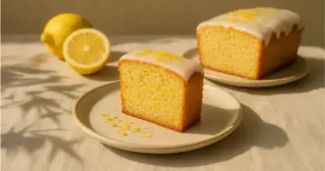 Ein Stück saftiger, veganer Zitronenkuchen mit Zuckerguss und Zitronenschale bestreut. Daneben liegen frische Zitronen.