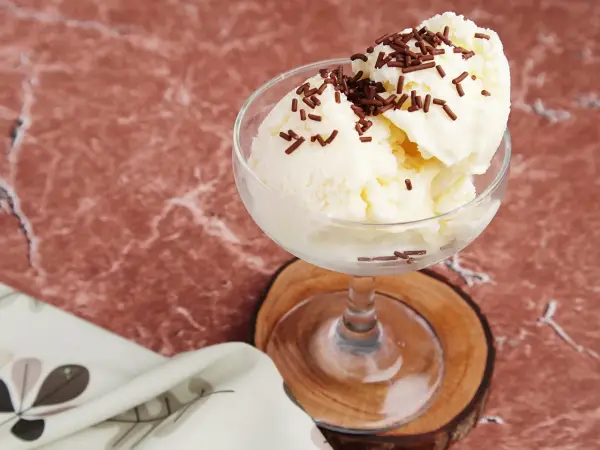Cremiges Litschi Eis mit Schokostreuseln in einem Glas auf einem Holzuntersetzer.