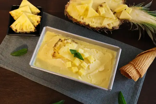 Erfrischendes Ananas Eis in einer Metallform, garniert mit Ananasstücken und Minze. Daneben liegen Ananasspalten und eine Waffel.