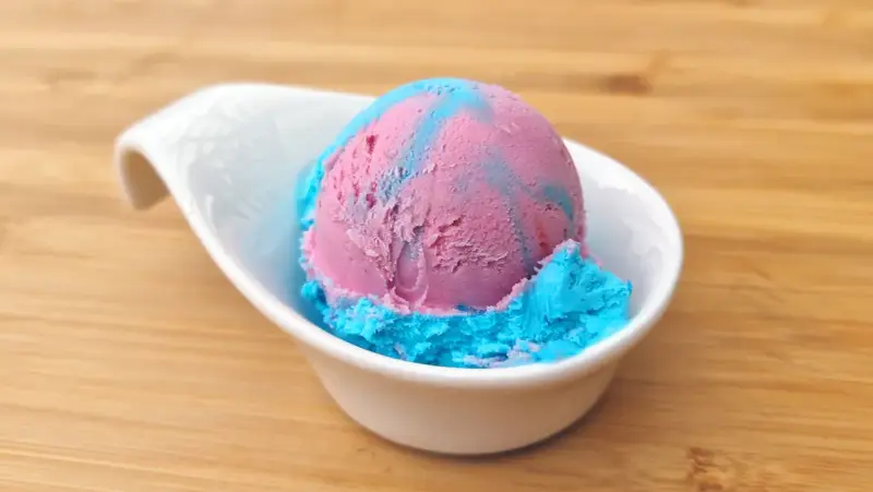 Ein Löffel eis mit kaugummi Geschmack, in rosa und blau, serviert in einer weißen Schale auf Holzuntergrund.