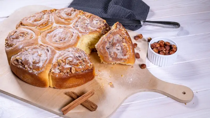 Ein köstliches Zimtschnecken Rezept: goldbraune, mit Nüssen und Puderzucker bestreute Schnecken auf einem Holzbrett, daneben Zimtstangen und Haselnüsse.
