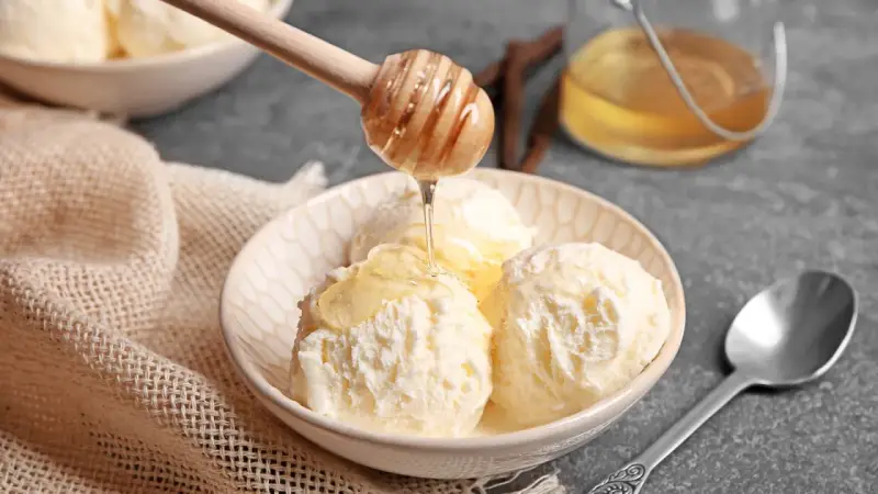 Schneeballgroße Kugeln Vanilleeis werden mit flüssigem Honig beträufelt. Ein Holzlöffel tropft goldenen Honig auf das cremige Eis.